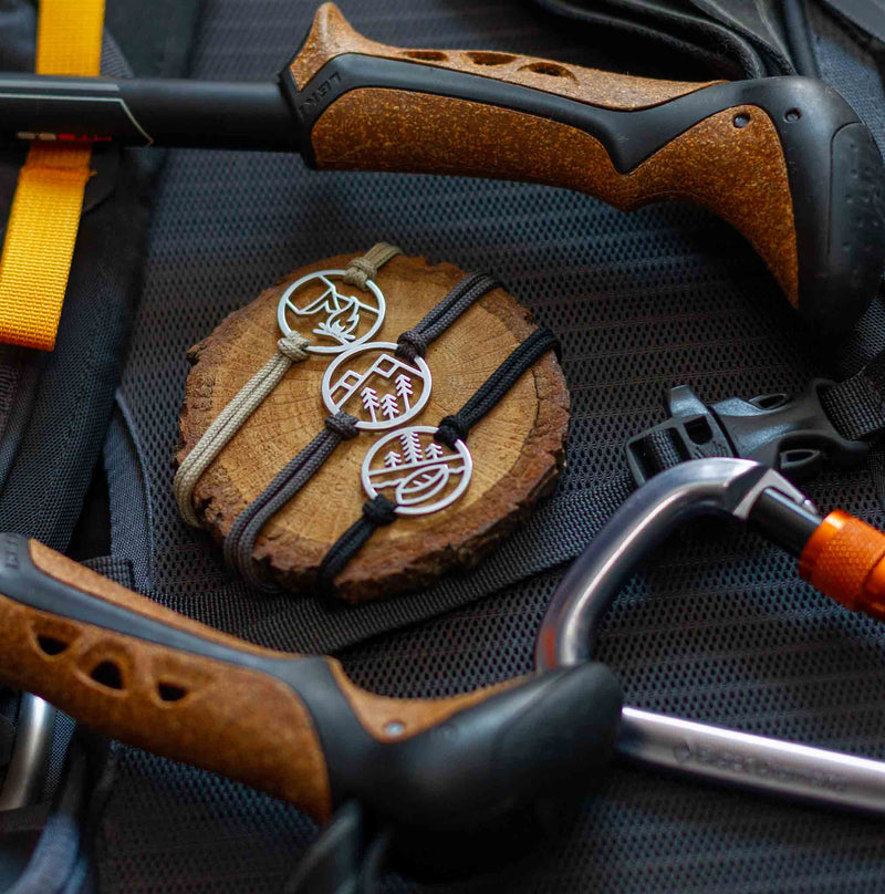 three tarma happy camper collection bracelets laid on a wood coaster that is sitting the back of a backpacking pack and surrounded by the handles of hiking poles and a carabiner